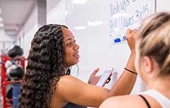 Two female students working on white board with exercise equipment behind them.