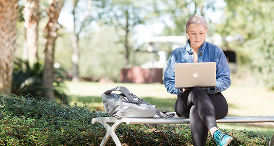 Student working on laptop sitting on a bench outside.