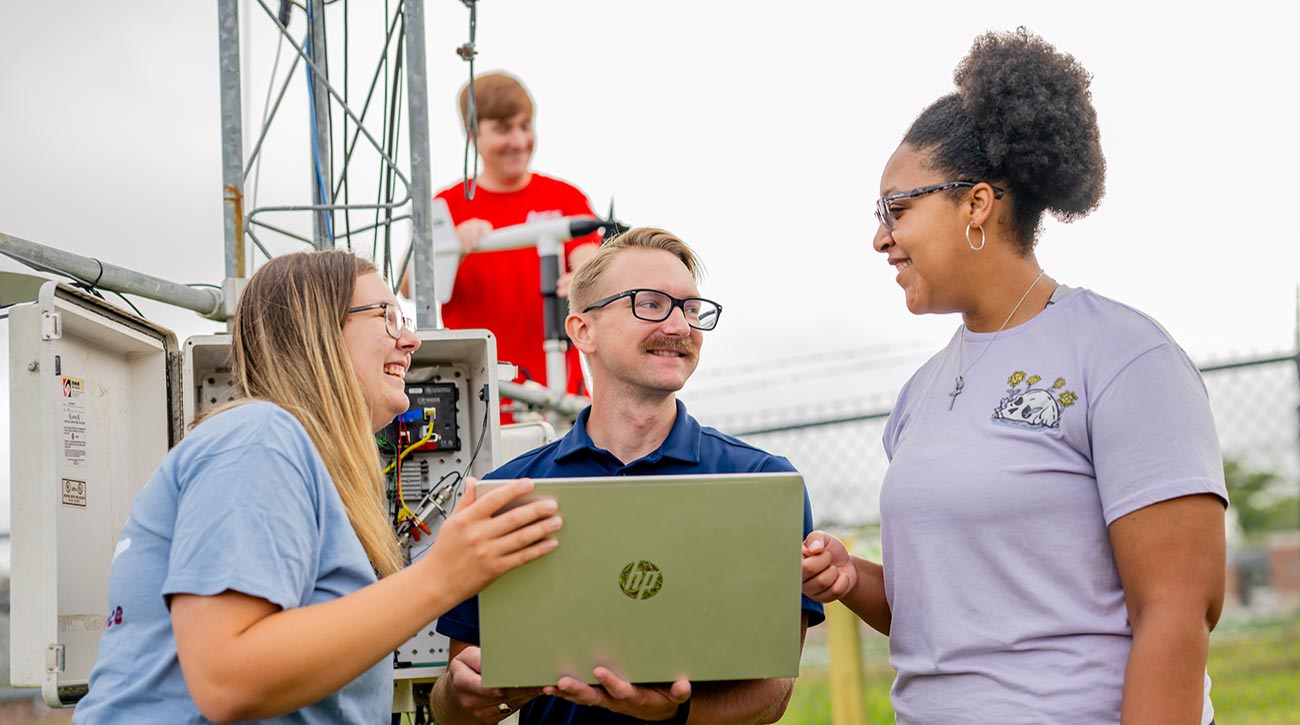Three students working with laptop outside tower.